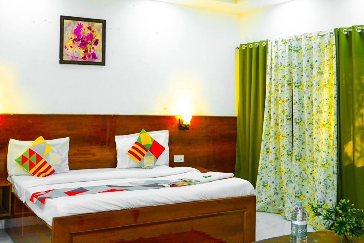 Une chambre d’hôtel avec un lit en bois, une petite table sur laquelle se trouve une bouteille, un tapis décoratif et un ventilateur de plafond.