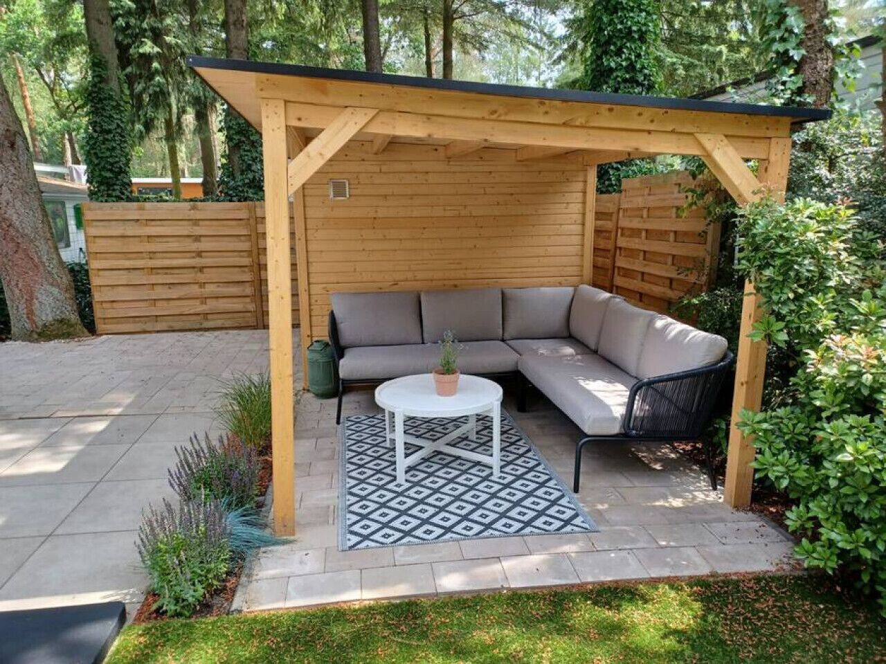 A covered outdoor seating area with a wooden pergola, a gray sofa, and a small round table with a potted plant.
