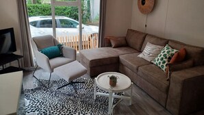 A modern living room with a sofa, armchair, and a small table. A television is on a stand to the left. - Cottage with a large garden (Hoenderloo)