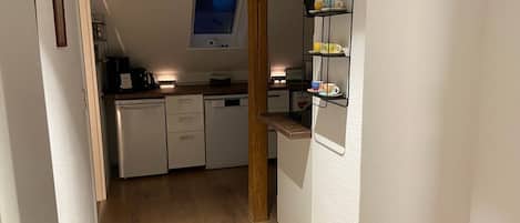 Een smalle gang die leidt naar een keuken met witte kasten, een vaatwasser en een raam boven de gootsteen.