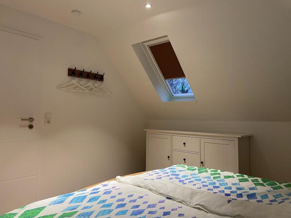 A bedroom with a skylight, a bed with a patterned bedspread, a wardrobe, and a small shelf with decorative items. - Holiday apartment Galgenweg (Friedeburg)
