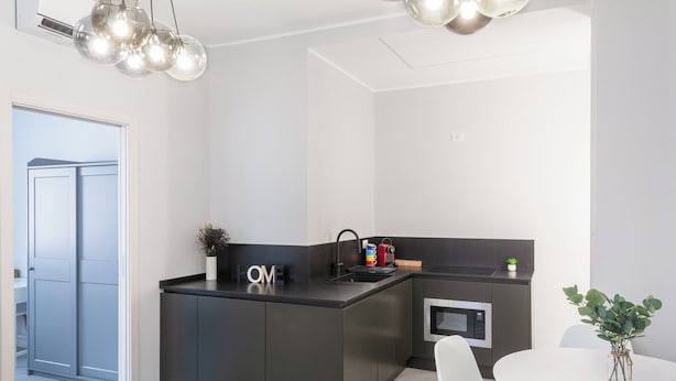 Uma cozinha moderna com bancada preta, micro-ondas embutido e uma pia pequena. Há uma mesa de jantar branca com cadeiras e um vaso de plantas.