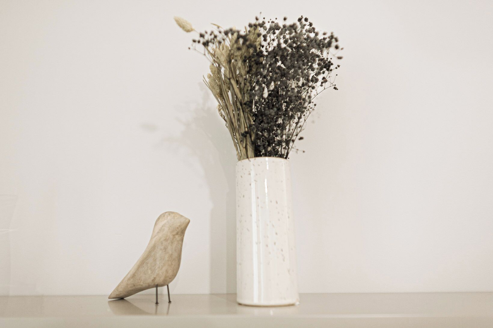 A white vase with a speckled pattern containing dried flowers and a wooden bird sculpture on a flat surface against a plain background.