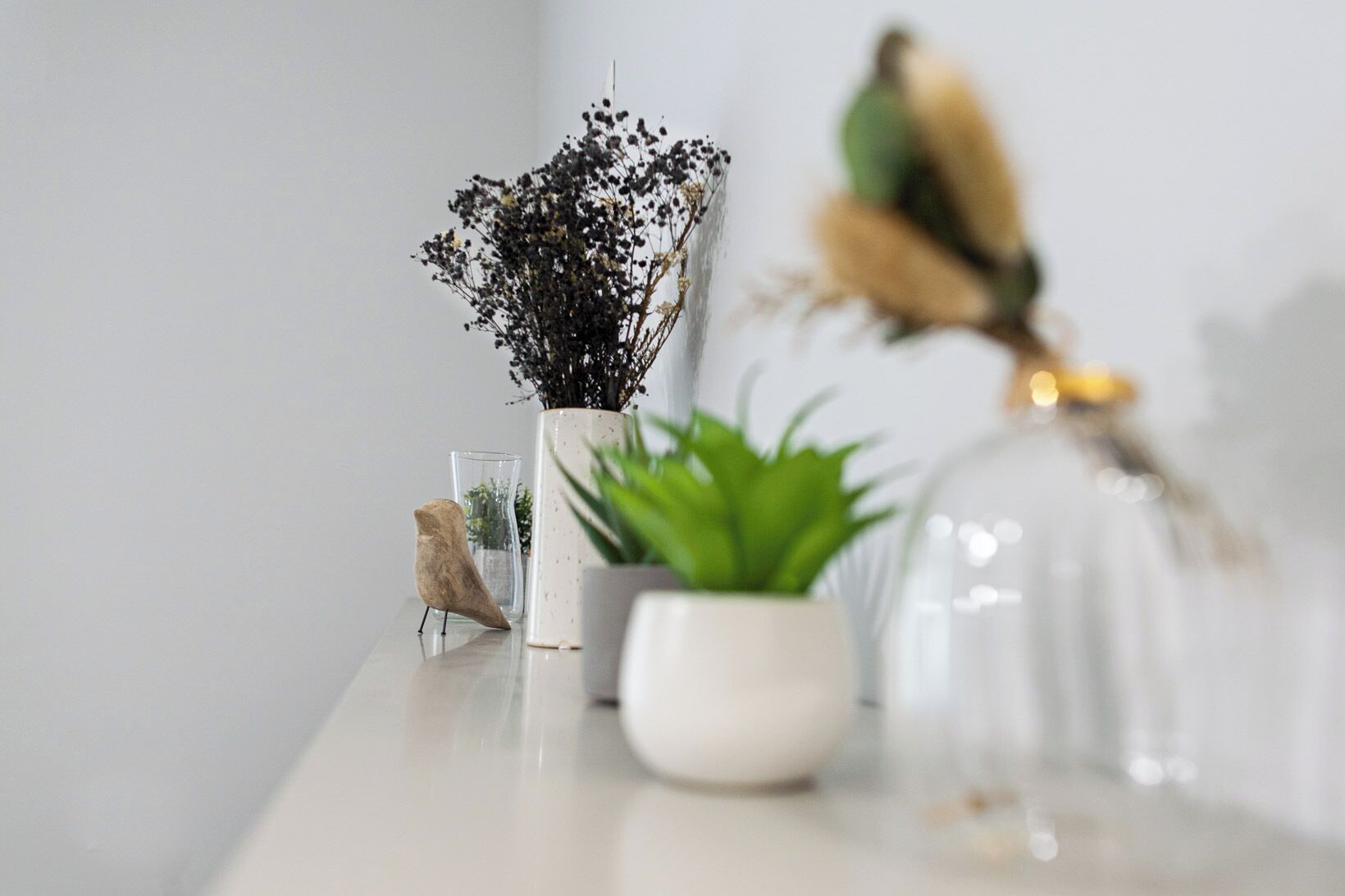 A white table with a potted plant, a glass vase with dried flowers, and a clear glass jar with a decorative item.