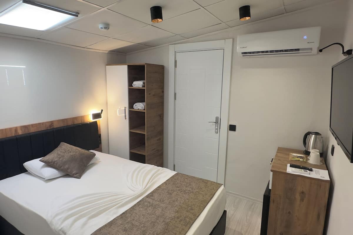 A hotel room with a bed, a wooden wardrobe, a small desk with a kettle, and a TV.