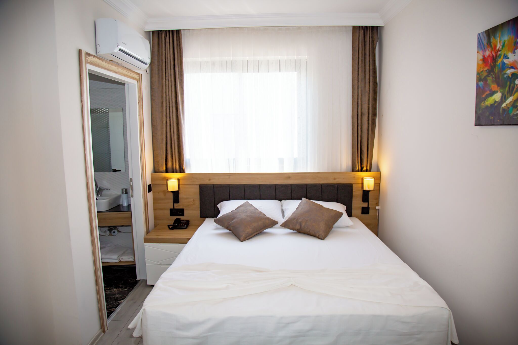 A hotel room with a bed, bedside lamps, a window with curtains, a painting on the wall, and an air conditioning unit.