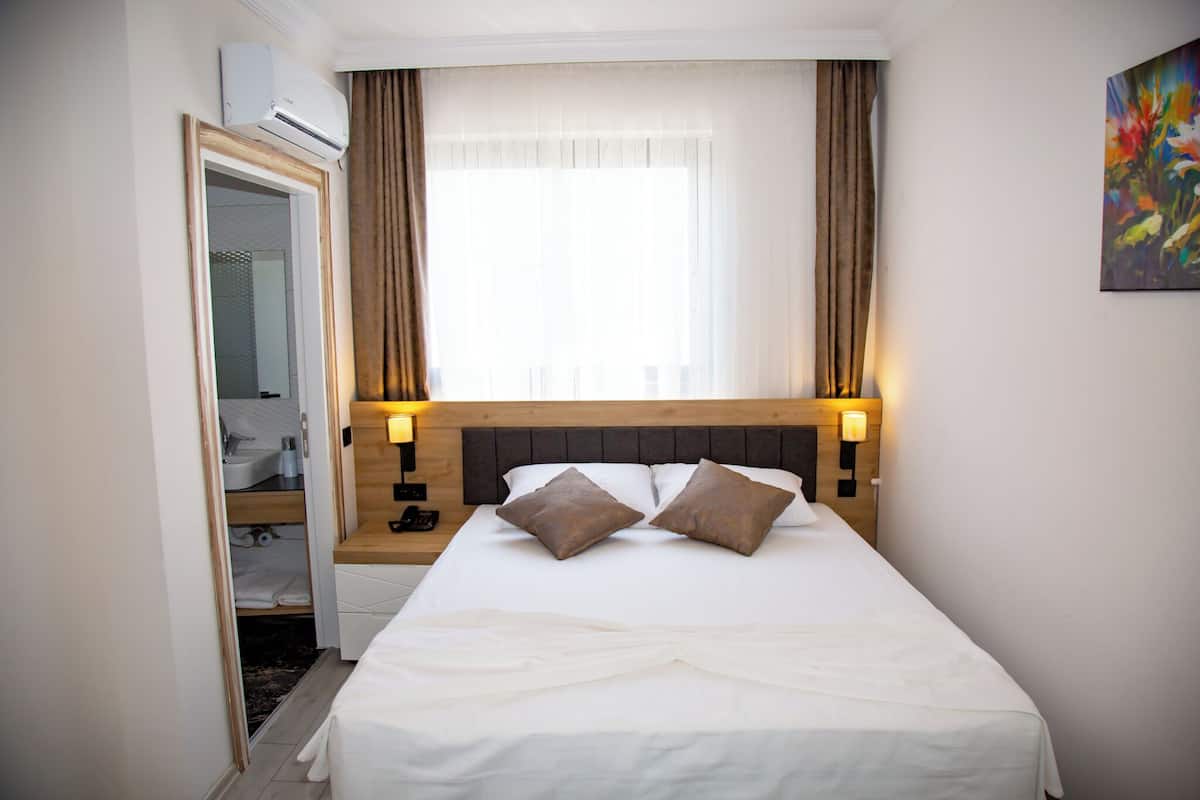 A hotel room with a bed, bedside lamps, a window with curtains, a painting on the wall, and an air conditioning unit.