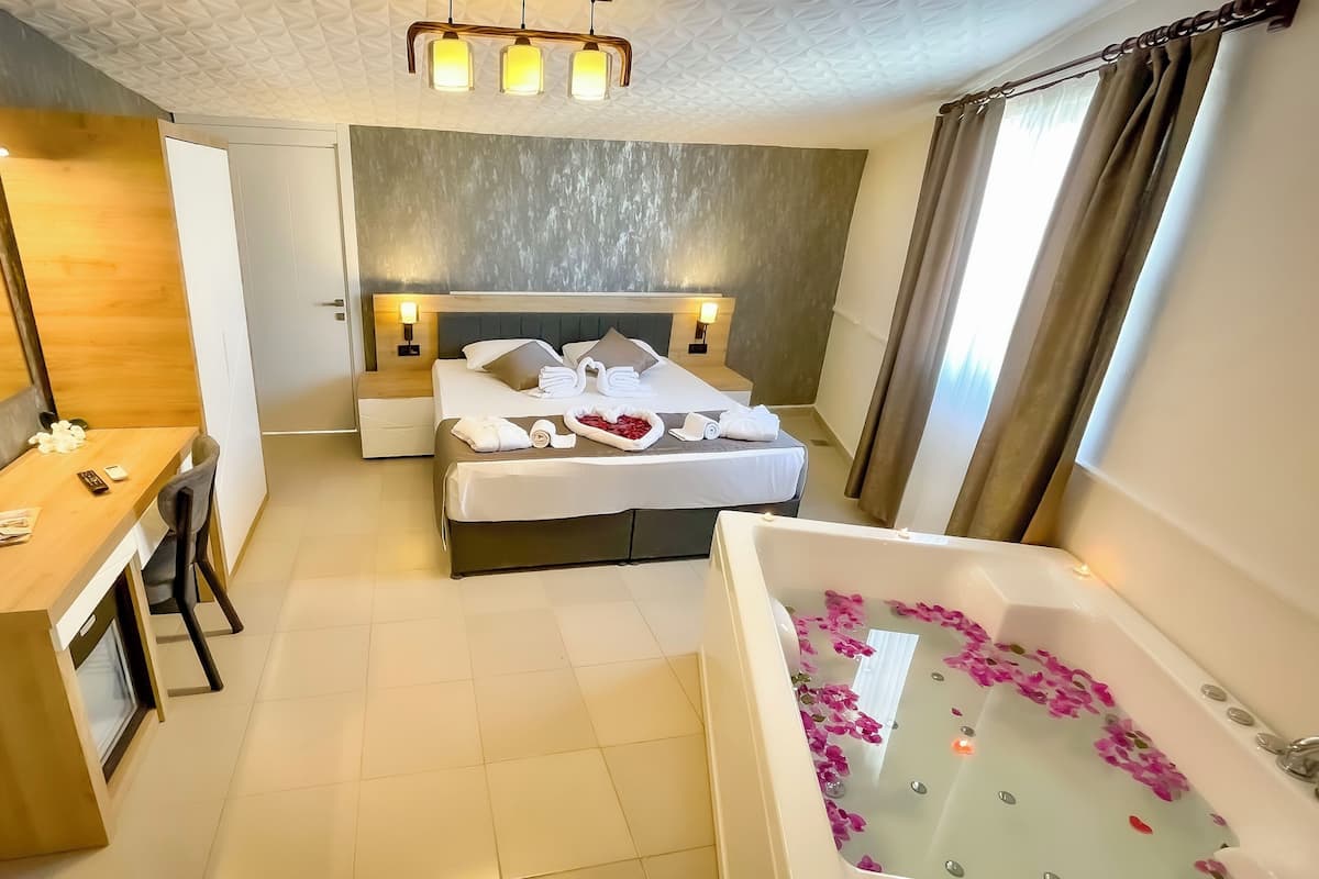 A hotel room with a bed, a desk, a chair, a bathtub with rose petals, and a ceiling light fixture.