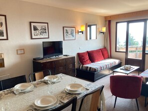 A hotel room with a dining table set for four, a sofa with red cushions, a TV on a wooden cabinet, and framed pictures on the walls. - Apartment for 6/7 pers, mountain view, small pets welcome (Avoriaz)