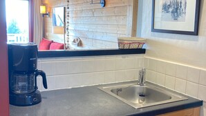 Una cocina con cafetera, un lavabo, una ventana con cortinas y un cuadro en la pared.