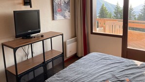 Una habitación de hotel con cama, televisor sobre un escritorio, un cuadro y ventana con vista a la montaña.