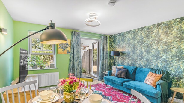 Ein Wohnzimmer mit einem blauen Sofa, einem Esstisch gedeckt mit Tellern und Gläsern sowie einem farbenfrohen Teppich.