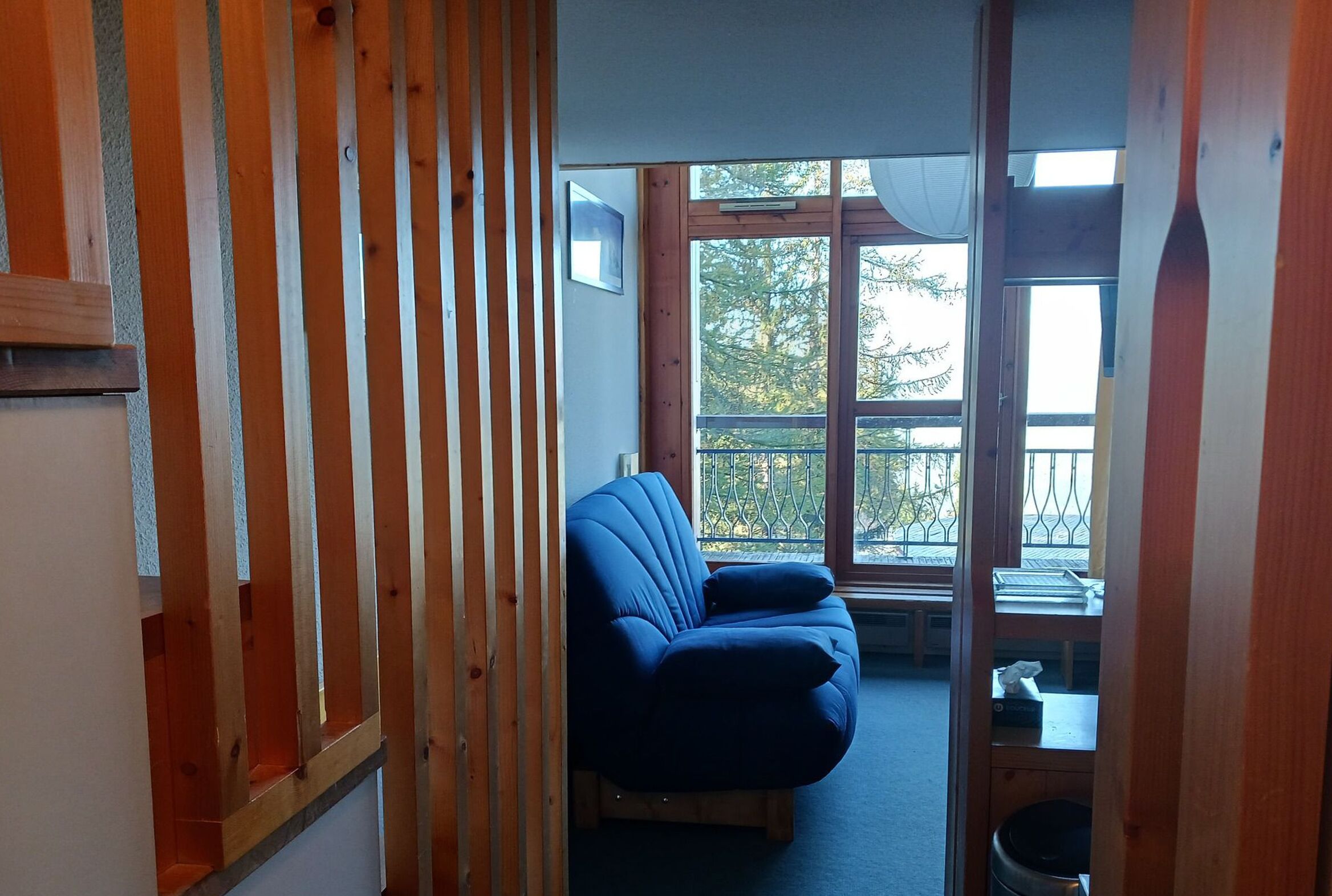 Une pièce avec un canapé bleu, des lambris en bois, une fenêtre donnant sur des arbres et une petite table sur laquelle sont posés divers objets.