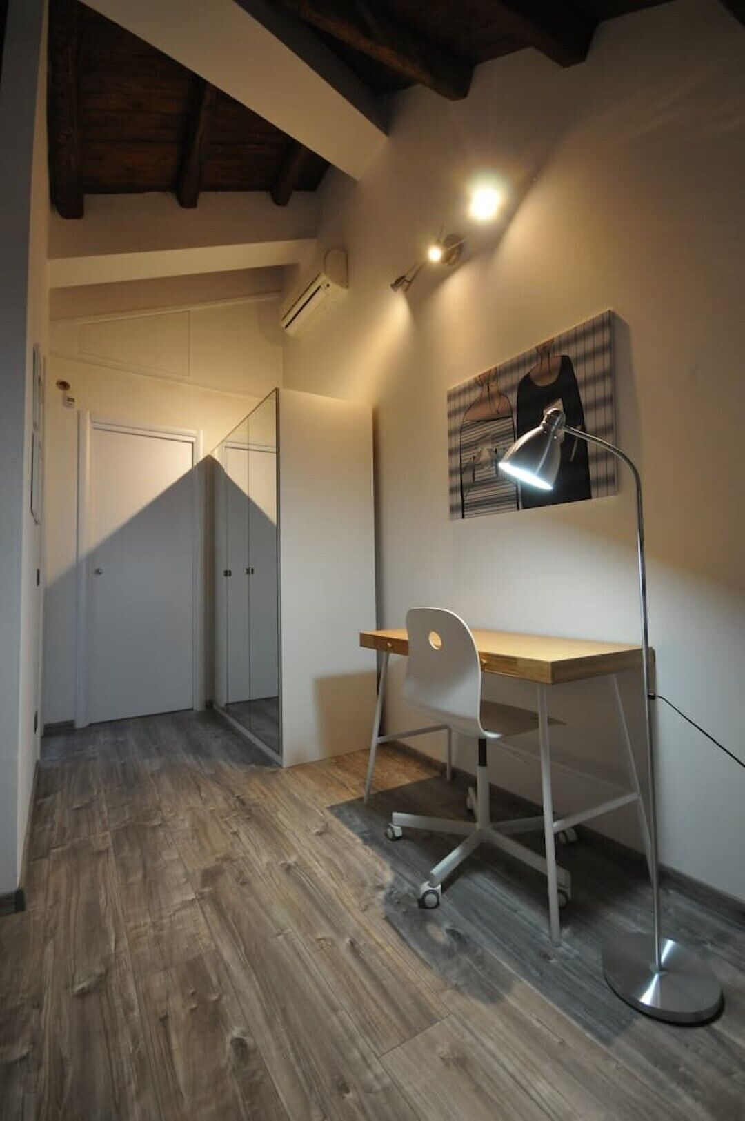 A room with a wooden desk, a white chair, a floor lamp, and a painting on the wall.