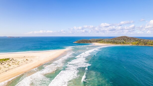Un paysage côtier avec une plage de sable, des eaux bleues et limpides, et un terrain vallonné en arrière-plan.