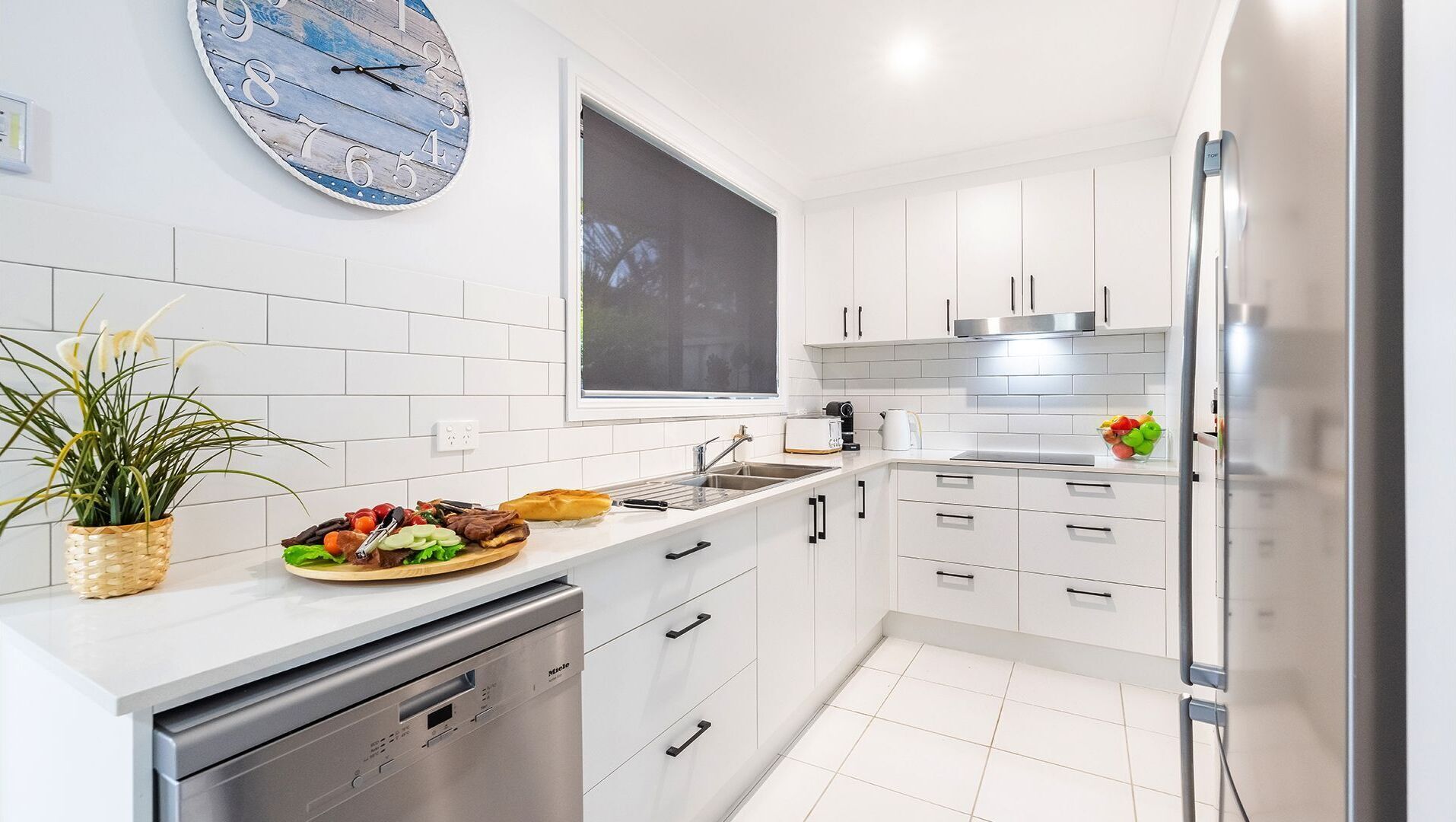A modern kitchen with white cabinetry, a built-in dishwasher, a large window, and a decorative clock.