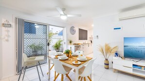 Une cuisine moderne avec des meubles blancs, un coin repas avec des chaises blanches et une table en bois, et un grand téléviseur à écran plat posé sur un meuble télé blanc.