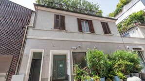 A two-story building with a grey facade, brown shutters, and a small entrance with steps. There are potted plants and a white sculpture in front. - BnButler - Archimede 53 (Milan)