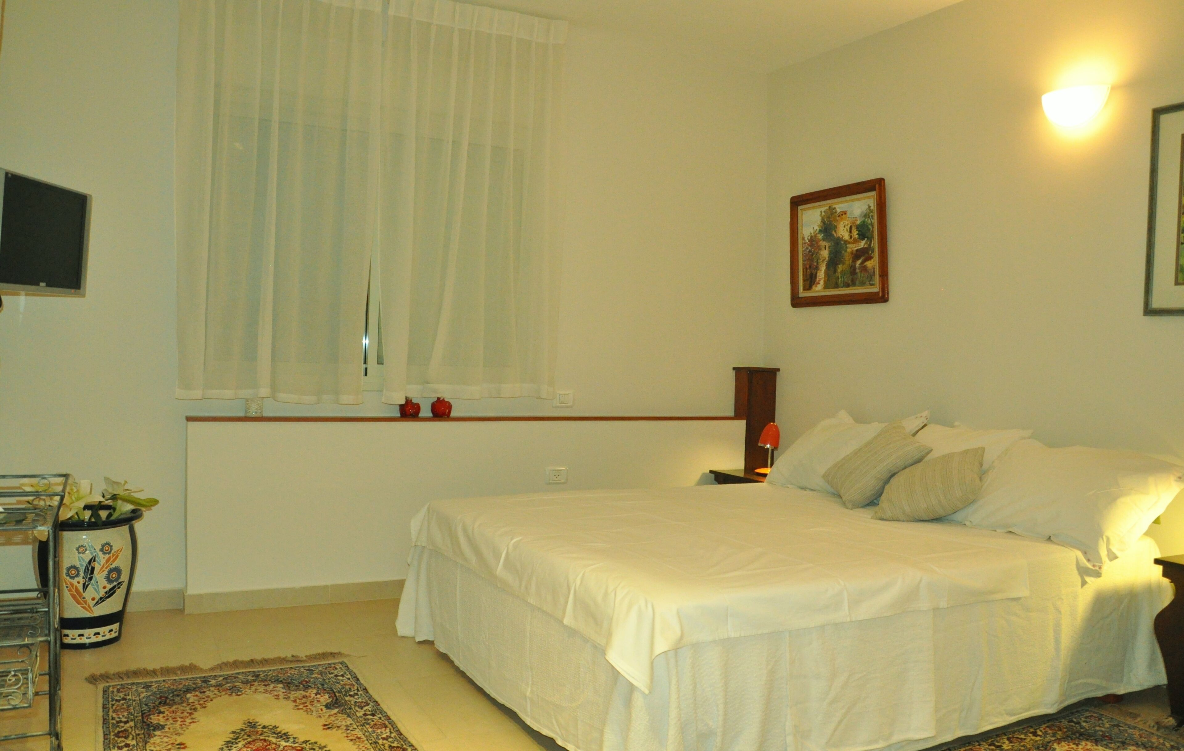 Une chambre à coucher avec un lit, une fenêtre avec des rideaux légers, une table de chevet avec une lampe et un téléviseur fixé au mur.