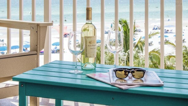 Une table avec une bouteille de vin, deux verres et une paire de lunettes de soleil, sur un balcon donnant sur une plage.