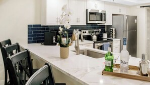 Une cuisine moderne avec des meubles blancs, un réfrigérateur en acier inoxydable, un four à micro-ondes et une bouilloire. On y trouve également un coin repas avec des chaises noires et une table blanche.