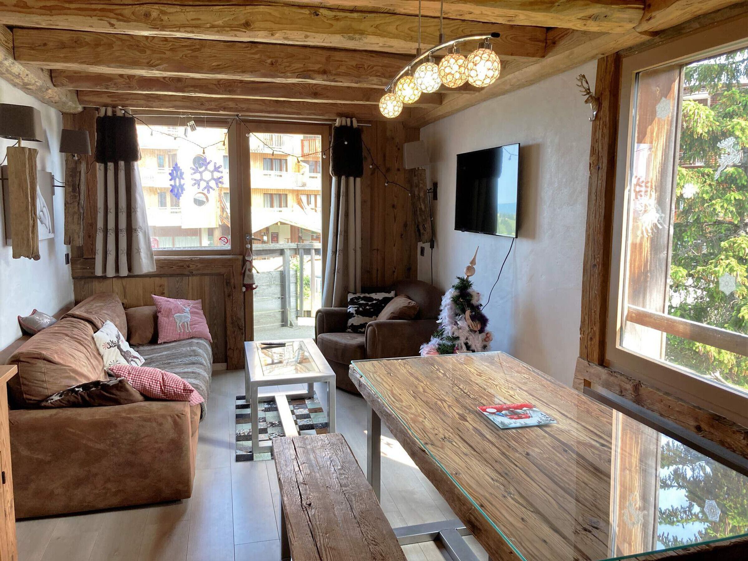 A cozy cabin interior with a wooden ceiling, a sofa, a coffee table, a dining table, and a TV mounted on the wall.