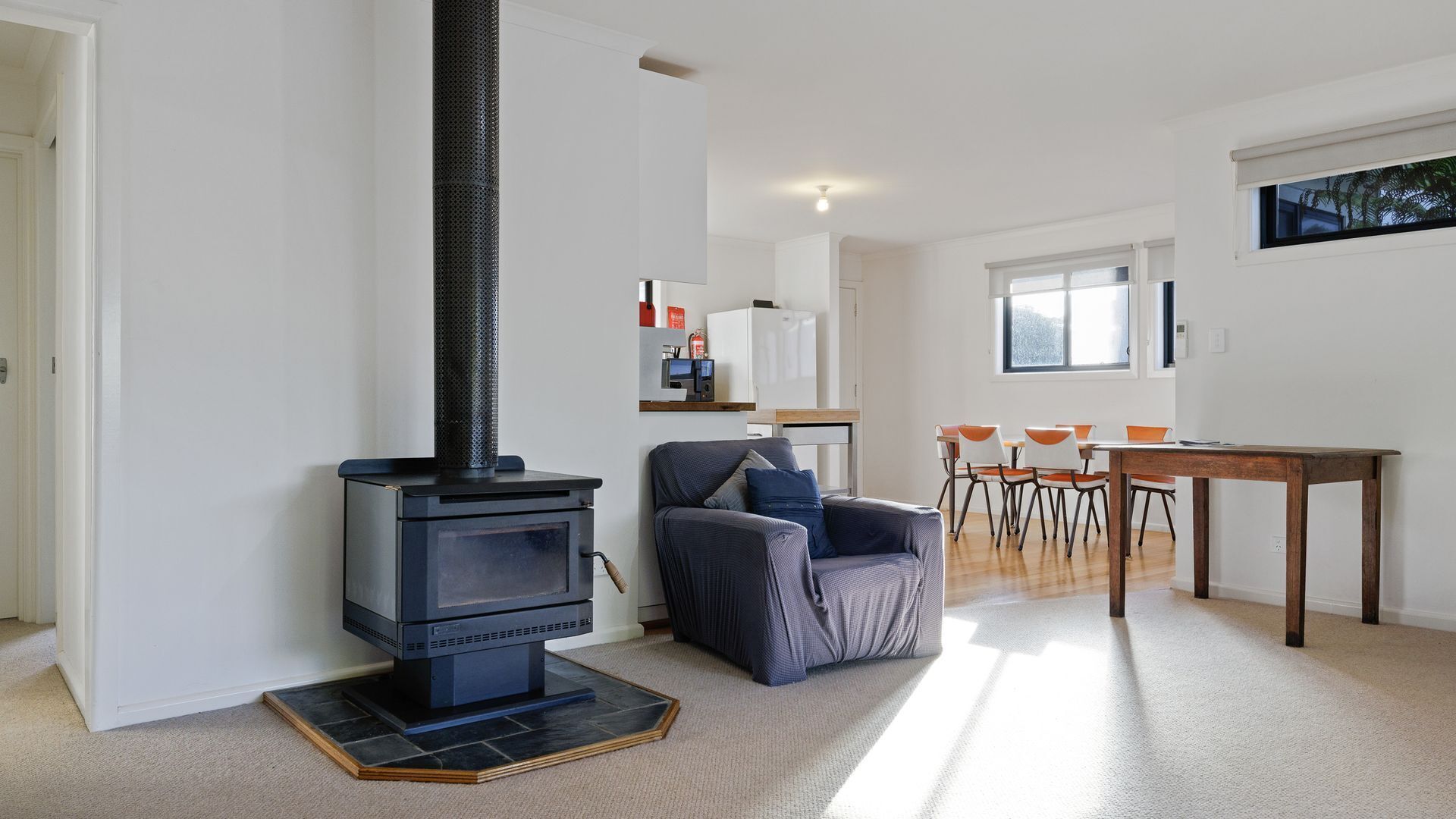 A modern living room with a black wood-burning stove, a grey armchair, a dining table with orange chairs, and a kitchen area in the background.