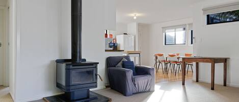 A modern living room with a black wood-burning stove, a grey armchair, a dining table with orange chairs, and a kitchen area in the background.