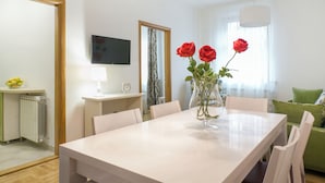 A modern dining area with a white table, four chairs, a vase with red roses, a television, a mirror, and a green sofa. - Apartments IvanBeg (Belgrade)