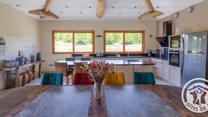 A modern kitchen with a central island, wooden beams, large windows, and a dining area with colorful chairs.