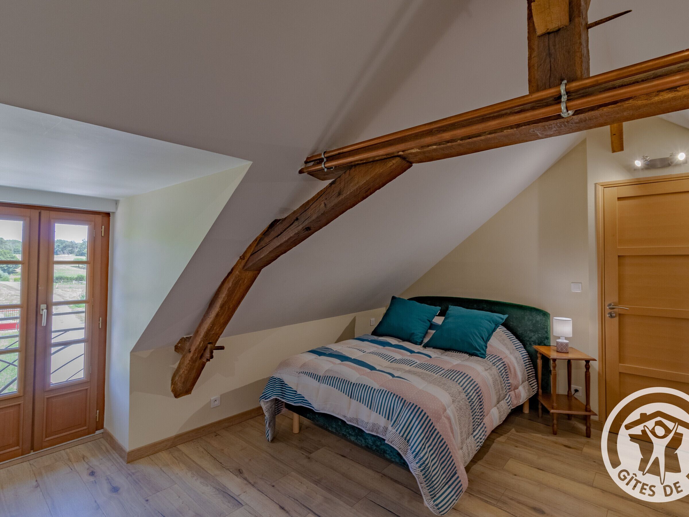 A bedroom with a sloped ceiling, a bed with teal and white bedding, a wooden headboard, and a small bedside table with a lamp.