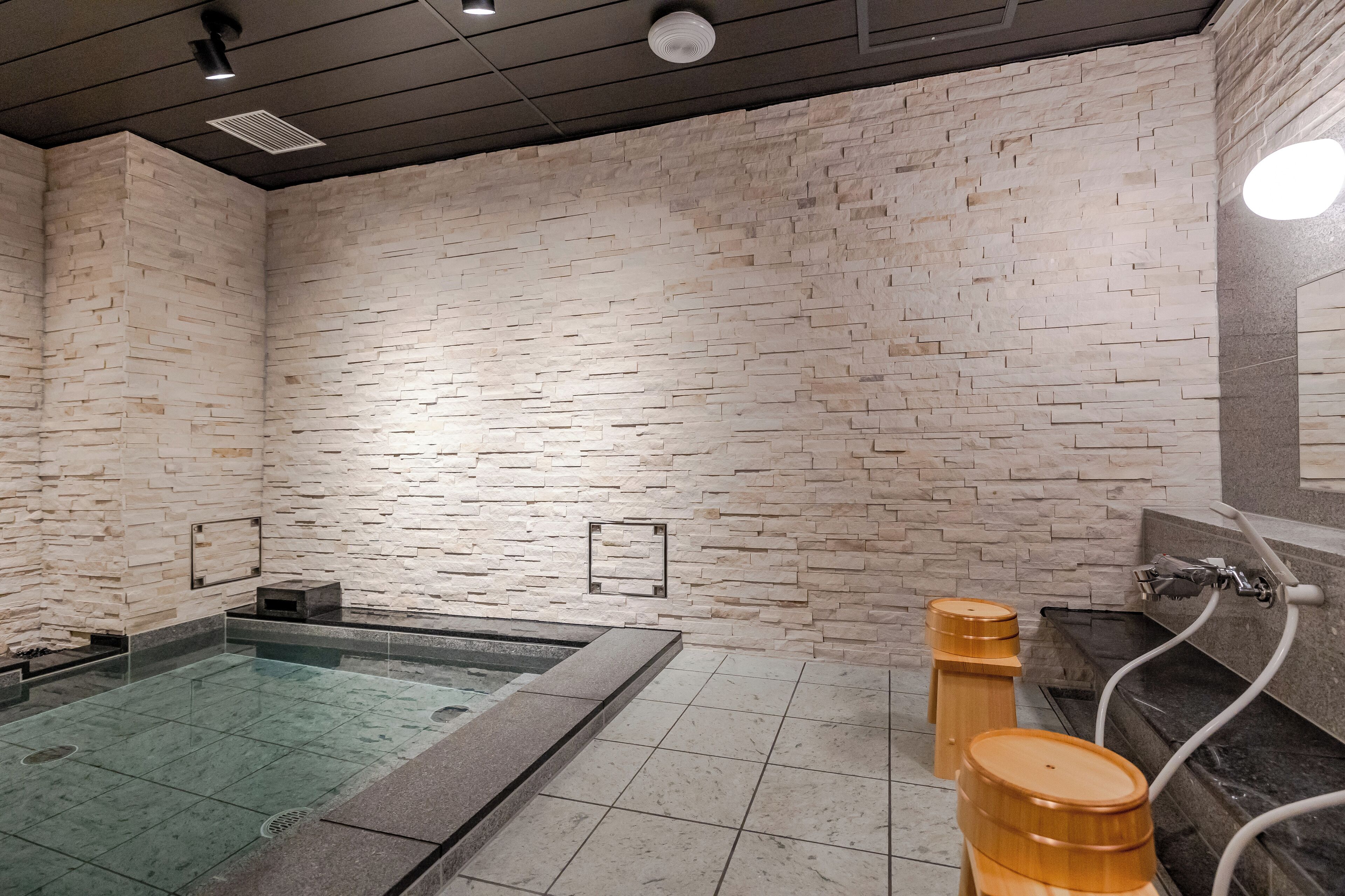 A modern spa area with a stone wall, a glass-enclosed hot tub, and wooden barrels.