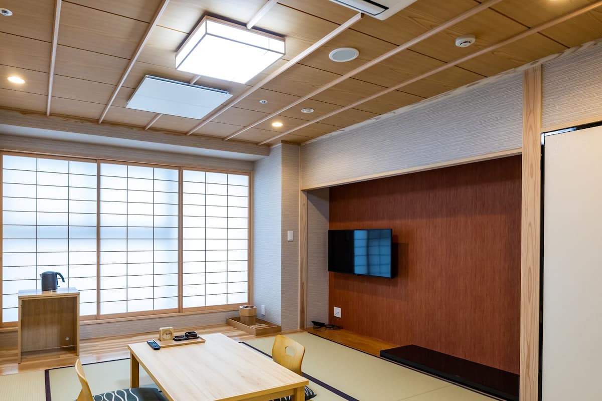 Une pièce de style japonais traditionnel, avec un sol recouvert de tatamis, une petite table basse avec un coussin, un téléviseur fixé à un mur en bois et une grande fenêtre à panneaux coulissants de type shoji.