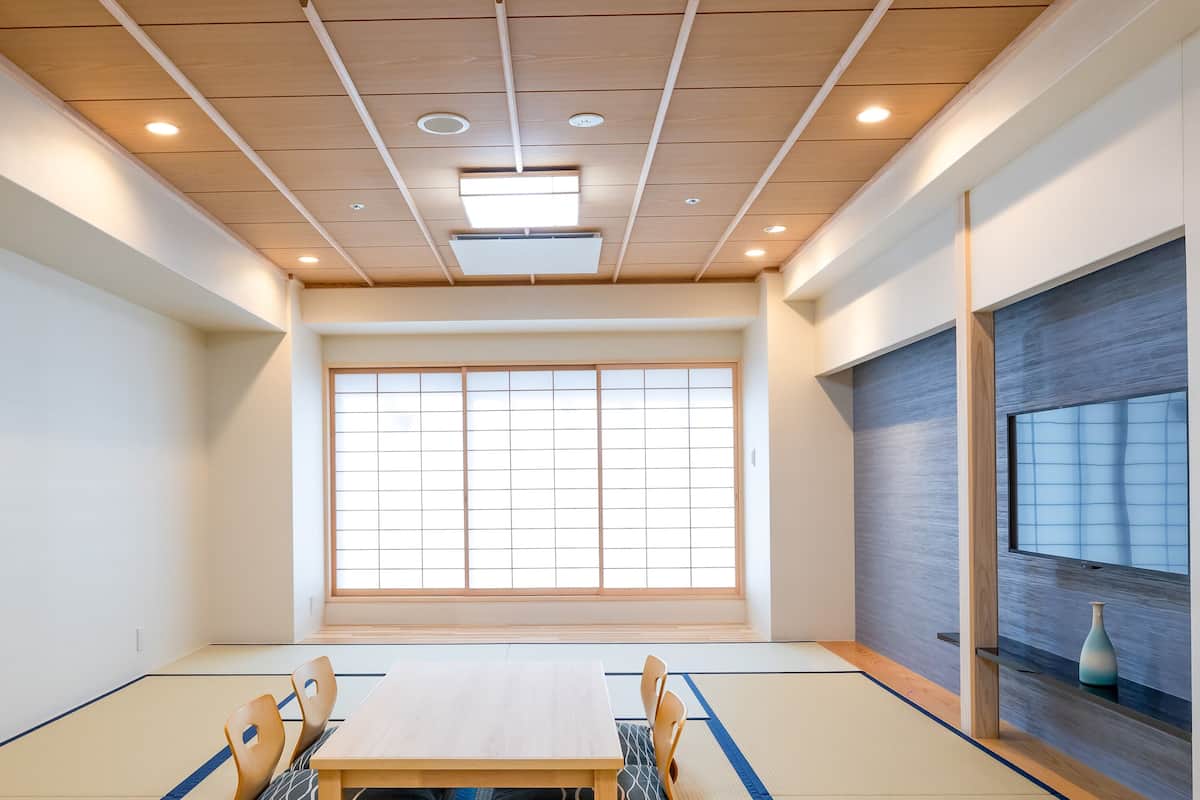 Une salle de réunion avec une table en bois, des tatamis, une grande fenêtre à panneaux coulissants de style shoji et un vase posé sur une étagère.