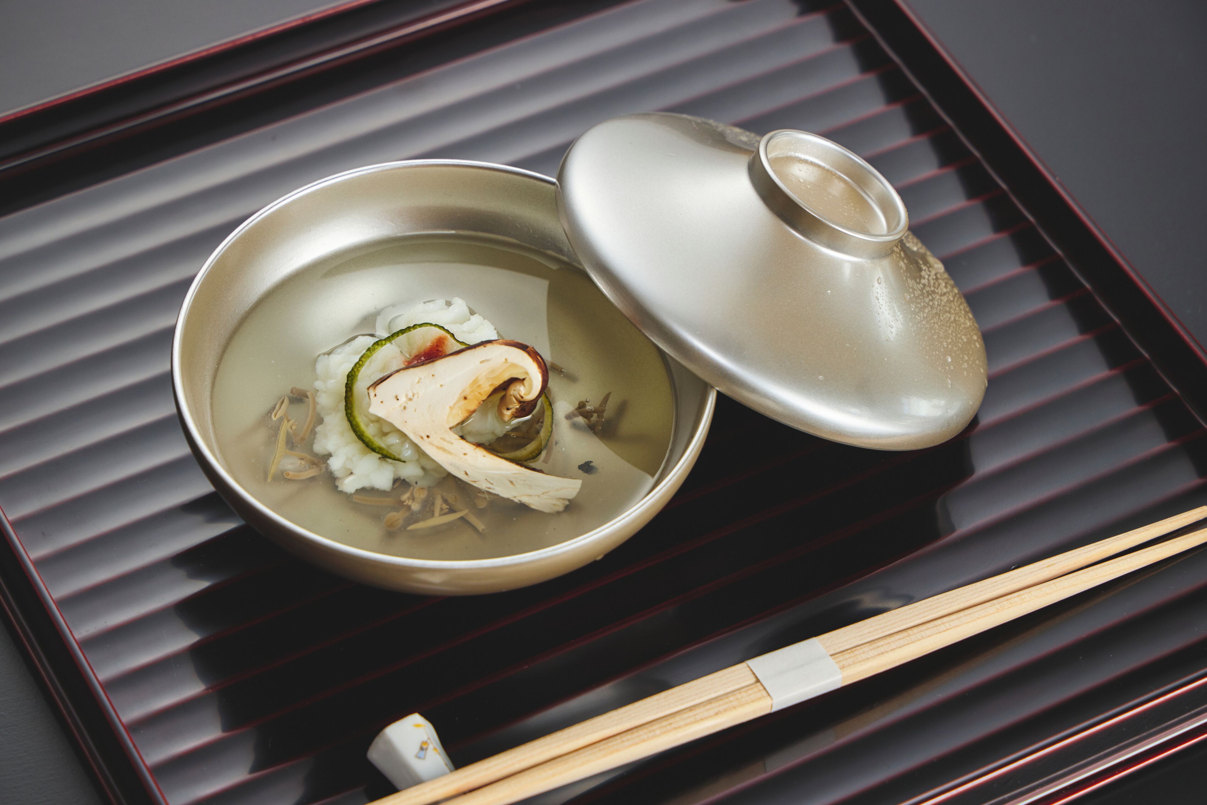 A bowl of soup with chopsticks and a lid on a tray.