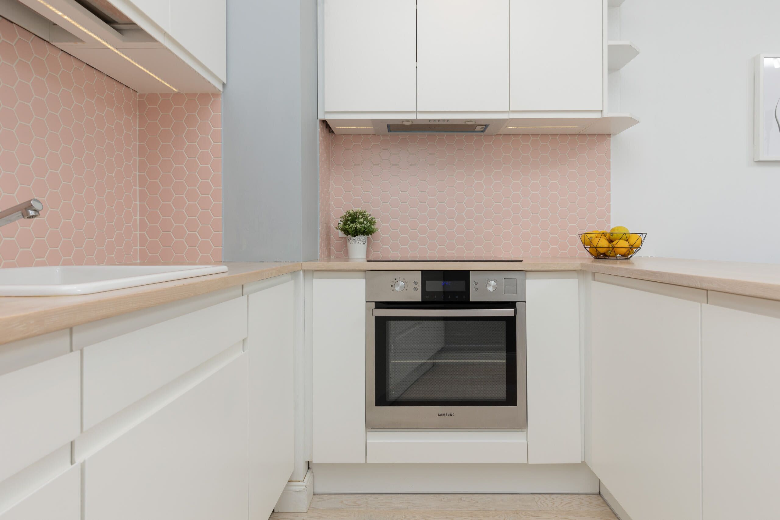 A compact kitchen with white cabinets, a stainless steel oven, a small potted plant, and a bowl of lemons on the countertop.