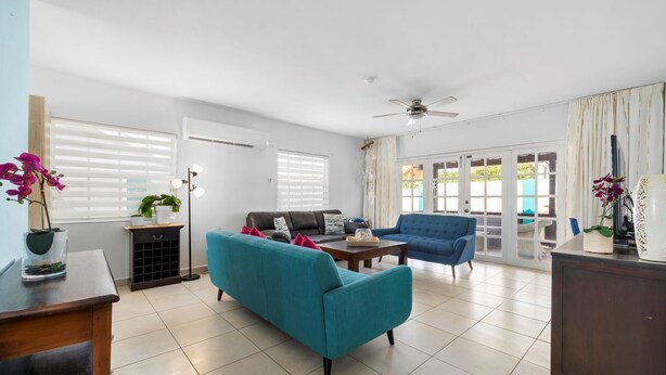 A spacious living room with a teal sofa, a blue armchair, and a wooden coffee table. There is a ceiling fan, a TV, and a large window with blinds. - Cozy home with private pool! Close to everything! Great dining options! Palm Beach and Hadicurari Beach! (Noord)