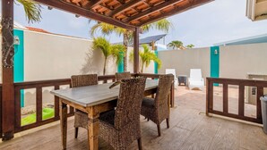 An outdoor patio with a wooden table and wicker chairs, surrounded by a wooden railing and palm trees. - Cozy home with private pool! Close to everything! Great dining options! Palm Beach and Hadicurari Beach! (Noord)