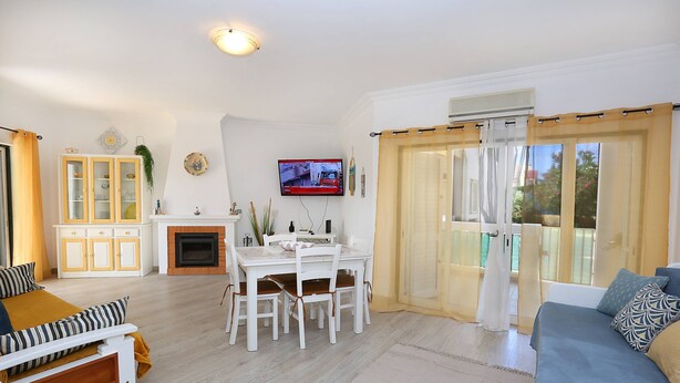 A living room with a sofa, a dining table with chairs, a TV, a fireplace, and a view through the curtains. - Apartment in Condominium with Swimming Pool (Alvor)