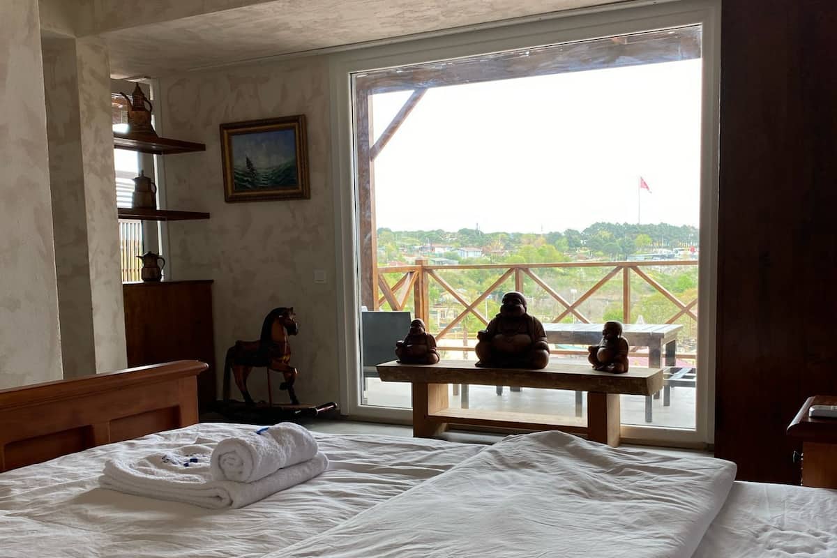A bedroom with a large bed, a wooden headboard, a wooden nightstand, a small table with a statue, and a window with a view of a balcony and buildings.