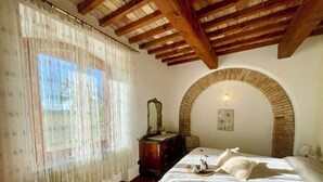 A bedroom with a wooden ceiling, a brick archway, a bed with a white bedspread, a dresser, and a window with sheer curtains. - A magnificent Tuscan farmhouse, surrounded by greenery between the sea and the hills (Pomaia)