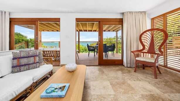 Una habitación moderna con suelo de madera, un sofá con cojines estampados, una mesa de madera con una revista y una silla de mimbre. Hay puertas correderas de vidrio que dan a una terraza exterior con vista al mar.