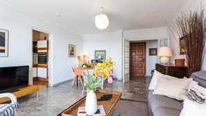 Un salon moderne avec un téléviseur à écran plat, un canapé, une table basse ornée d’un vase et de télécommandes, un coin repas avec une table et des chaises, et une cuisine en arrière-plan.