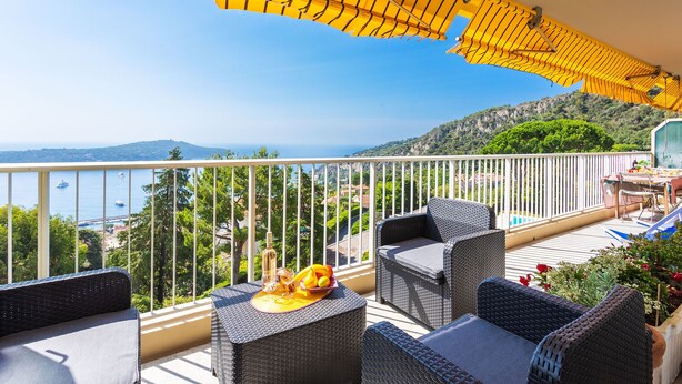 Un balcon agrémenté de meubles en osier, une table avec des fruits et des boissons, et une vue sur la mer et les collines.