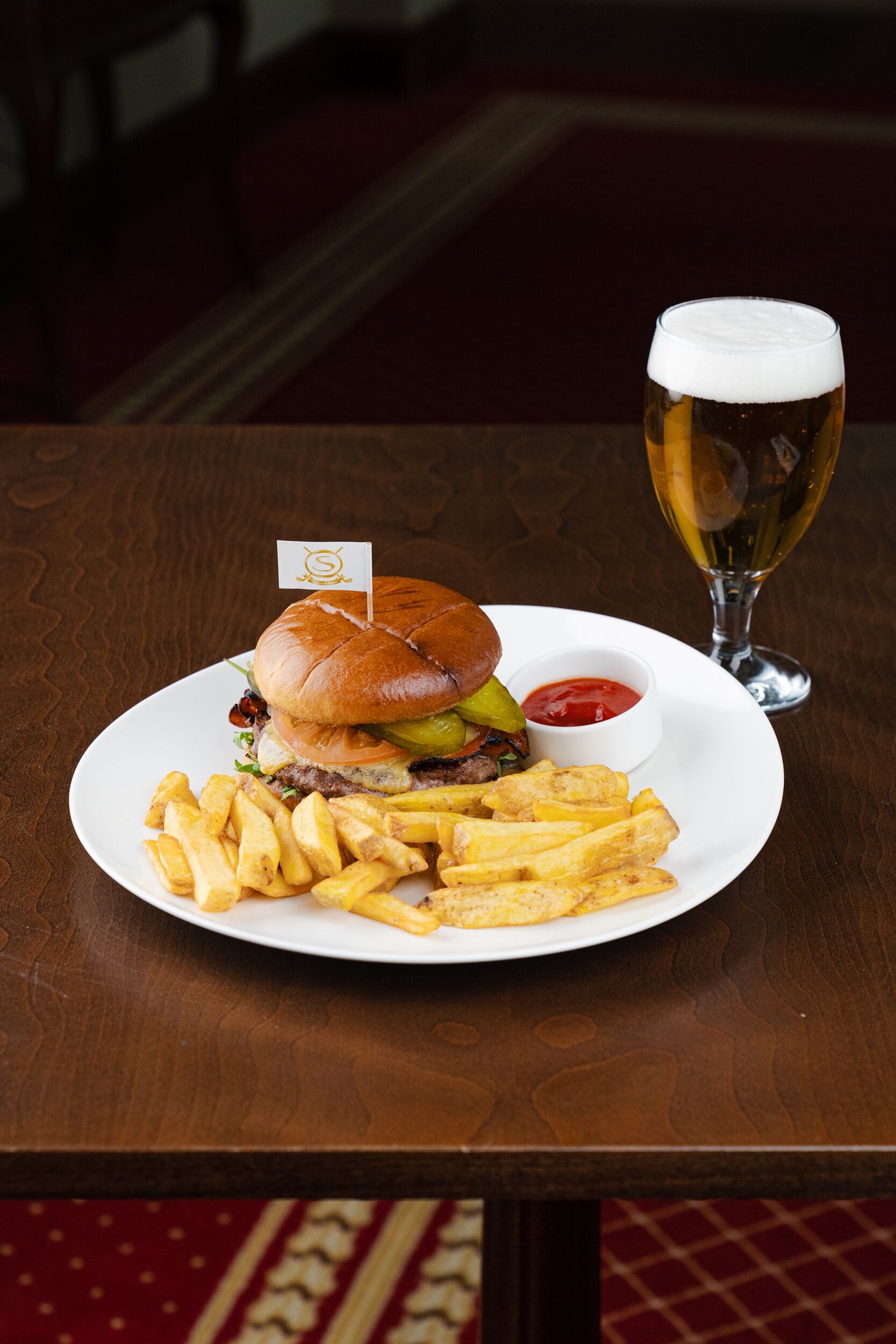 A plate with a burger, fries, and a glass of beer on a wooden table.