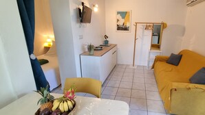 Un espacio habitable compacto con sofá, una mesa de comedor con fruta, una pequeña cocina y un televisor montado en la pared.
