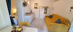 A compact living space with a sofa, a dining table with fruit, a small kitchen area, and a TV mounted on the wall.