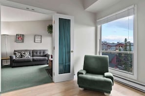 A modern living room with a grey sofa, a green armchair, a teal door, and a large window offering a city view.