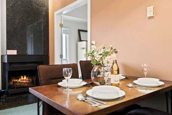 A dining table set with plates, glasses, and cutlery, a vase with flowers, and a bottle of wine, with a fireplace in the background. - Powder Pointe | Hot Tub & Ski In/Ski Out (Big White Mountain)