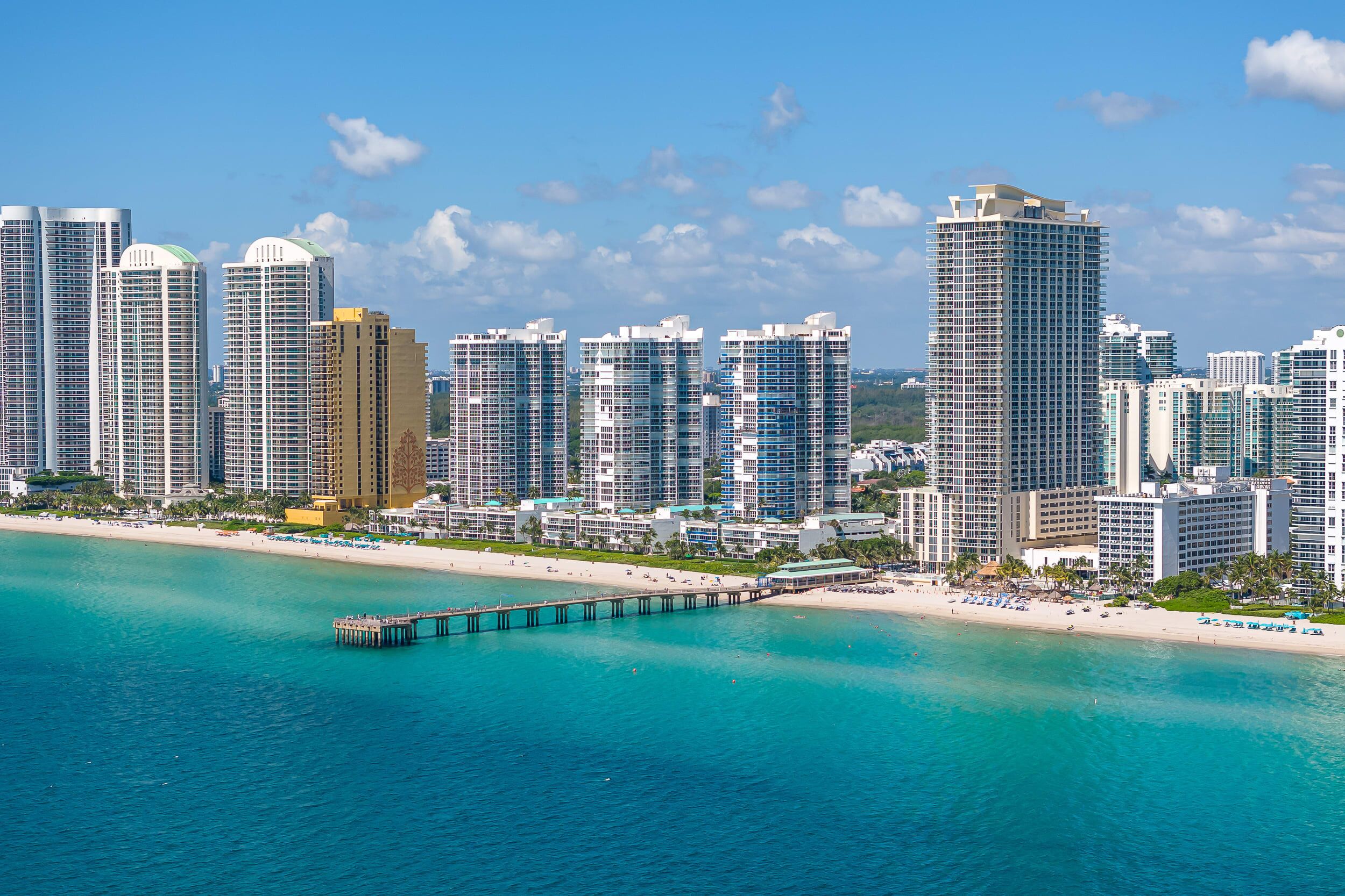 A coastal cityscape with high-rise buildings, a pier extending into turquoise waters, and a beach with umbrellas and sun loungers.